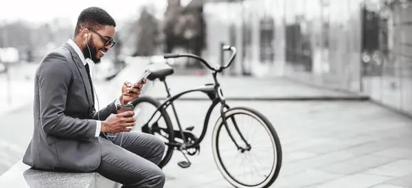 stock image An African American man is using his phone while sitting next to his bicycle. The setting is outdoors, providing a blend of work and leisure in a cityscape, web-banner