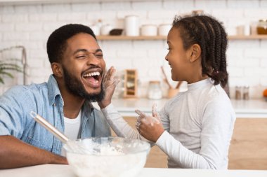 Afro-Amerikalı bir baba ve kızının mutfakta eğlenceli bir un savaşı yaparken yakalanan eğlenceli bir an neşe ve ebeveynliği simgeliyor.