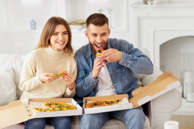 Bir adam ve bir kadın koltukta oturuyor, rahat bir ortamda pizza dilimlerinin tadını çıkarıyorlar..