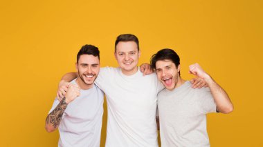 Üç neşeli arkadaş baş parmaklarıyla poz veriyor, gösterişli dostluk ve canlı sarı bir arka plana karşı mutluluk.