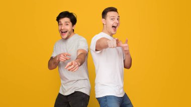 Canlı sarı bir arka planda kamerayı işaret eden iki neşeli genç adam, coşku ve neşe ifade ediyorlar.