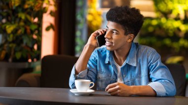 Genç bir Afro-Amerikan adam bir masada oturuyor, telefonuyla sohbet ediyor. Evde otururken odaklanmış ve dikkatli görünüyor..