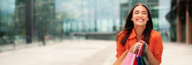 Alışveriş torbalarıyla poz veren, elinde renkli kâğıt paketler tutan, hafta sonu şehirde yürüyüş yapan ve şehir dışında ayakta duran mutlu kadın portresi. Ticaret ve indirimler, kopyalama alanı