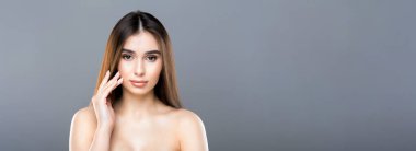 Uzun, kahverengi saçlı genç bir kadın, eli düz gri arka planda yüzüne dokunurken poz veriyor. Profesyonel stüdyo ışıkları altında sakin ve kendinden emin bir tavır sergiliyor..