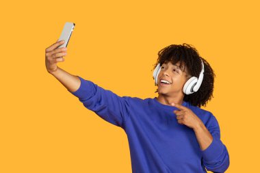 Kıvırcık saçlı genç bir adam sarı arka planda selfie çekiyor. O kulaklık takıyor ve mutlu bir şekilde gülümsüyor. İşaret parmağı kameraya doğrultulmuş, sanki baş parmağını kaldırıyormuş gibi..