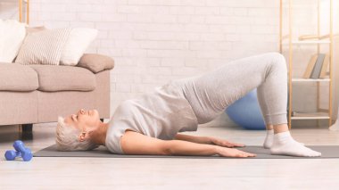 Kısa saçlı son sınıf öğrencisi bir kadın sırtüstü uzanmış geniş, aydınlık oturma odasında yoga minderinin üzerinde köprü egzersizi yapıyor. Rahat kıyafetler giyiyor ve yanında da bir çift dambıl var.
