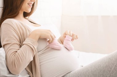 Hamile karnına bir çift pembe bebek ayakkabısı dayamış, neşeli, hamile bir anne yatakta oturuyor. Heyecan ve mutluluk saçıyor.