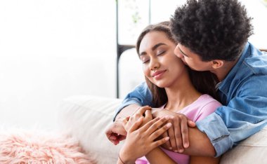 Kısa, kıvırcık siyah saçlı Afro-Amerikalı bir adam elini tutarken bir kızın yanağını öpüyor. Beyaz bir koltukta oturuyorlar, kopyalama alanı.