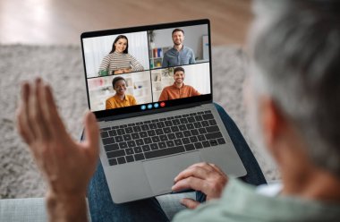 Üst düzey bir adam koltukta oturuyor, sırtı kameraya dönük, aktif olarak bilgisayarındaki bir video konferans görüşmesine katılıyor. Ekranda dört katılımcı var, hepsi de gülümsüyor ve sanal toplantıya katılıyorlar.
