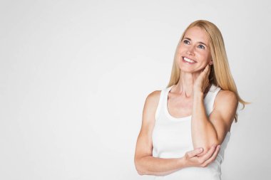 Beyaz atlet giyen ve yukarı bakan, sarı saçlı, gülümseyen bir kadının portresi. Kadınların arka planı sade, beyaz bir duvar..