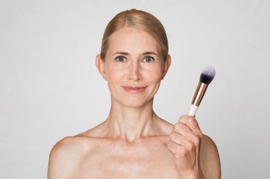 Sarışın ve açık tenli bir kadın beyaz arka plan önünde makyaj fırçası tutarken görülüyor. Doğrudan kameraya bakıyor ve hafifçe gülümsüyor..