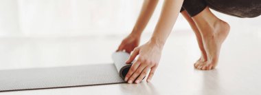 Bir kadın yoga minderini parlak ve havadar bir odada temiz, beyaz bir zemine yuvarlarken görülüyor. Atmosfer, yoga sonrası dinlendirici bir seans izlenimi uyandırıyor.