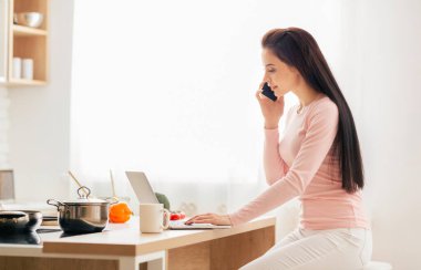 Bir kadın mutfak tezgahında oturmuş, telefonla konuşuyor. Etrafı çeşitli mutfak malzemeleriyle çevrili cep telefonuyla konuşurken odaklanmış görünüyor..