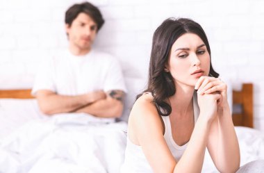 Görüntü, yatakta oturan üzgün ve düşüncelere dalmış bir kadını gösteriyor. Beyaz bir atlet giyiyor ve elleri önünde birbirine kenetlenmiş..