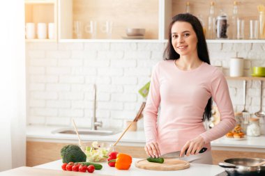 Bir kadın mutfakta durmuş, ahşap bir kesme tahtasındaki çeşitli renkli sebzeleri doğramaya odaklanmış. Bir elinde keskin bir bıçak tutuyor ve sebzeleri ustaca dilimliyor.