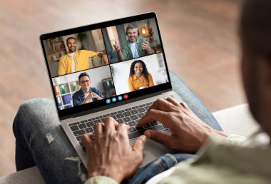 Kanepede oturan Afrikalı Amerikalı bir adam bilgisayarını kullanarak online bir video konferansına katılıyor. Laptop ekranında dört kişi görünüyor ve adam klavyede daktilo kullanıyor..