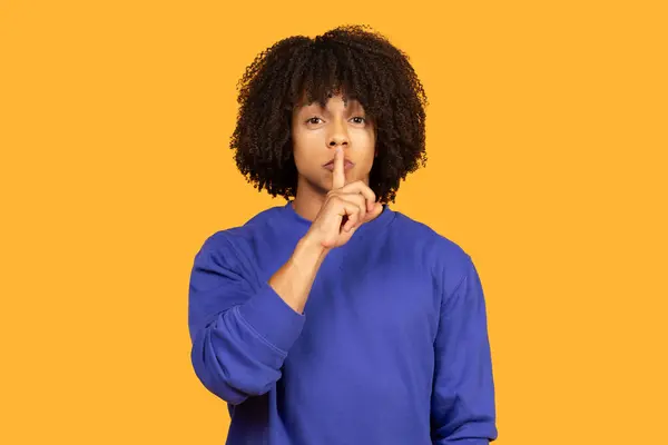 Stock image A man with curly black hair is wearing a blue sweatshirt and gesturing for silence with his finger to his lips against a yellow background.