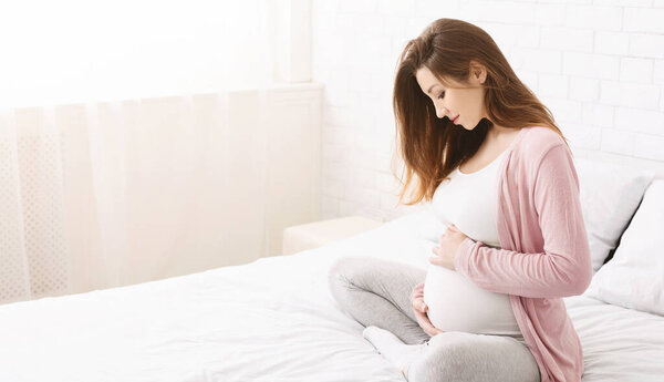A young pregnant woman is seated on a bed in a bright, minimalist bedroom during the daytime. She is gently cradling her belly and appears deep in thought, copy space