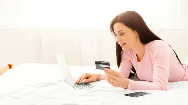 stock image A young woman lies on her bed in a white, bright bedroom. She is engaged in online shopping, holding her credit card while typing on her laptop. The morning sunlight filters through the window