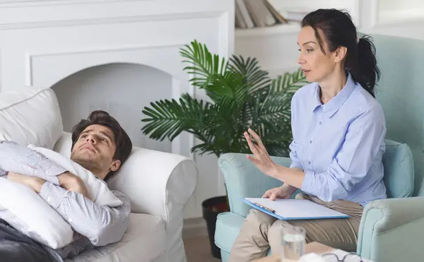 stock image A therapist engages with a man patient lying on a sofa, providing counseling in a contemporary living room with natural light and indoor plants.