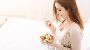 Bir kadın elinde bir kase salata tutarken görülüyor, sağlıklı bir yemek seçimi sergiliyor. Kasedeki renkli sebzeler ve sebzeler onun elinin ve giysisinin önünde duruyor.