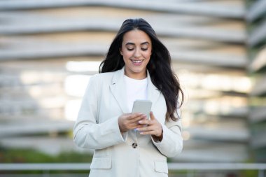 Beyaz ceketli ve beyaz atletli bir kadın modern bir şehir ortamında dururken akıllı telefonunu kullanıyor. Onun uzun, siyah saçları arkasında akıyor..