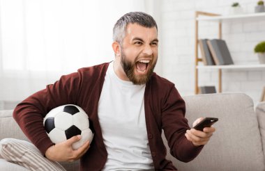 Sakallı bir adam futbol topunu tutarken televizyonda futbol maçını hevesle izler. Oturma odasındaki açık renkli bir kanepede oturuyor.
