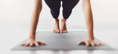 Tanımlanamayan bir kadın, kapalı bir yerde yoga minderinin üzerinde tahta bir pozisyonda görülüyor. Görüntü, gün boyunca iyi aydınlatılmış bir odada yoga egzersizi yapıldığını düşündüren bacaklara ve ellere odaklanıyor..