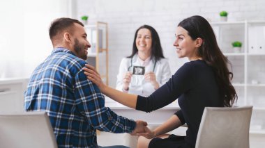 Bir çift, doktor muayenehanesinde yüz yüze oturup el ele tutuşuyorlar. Kadın gülümsüyor ve elini erkeğin omzuna koyuyor..