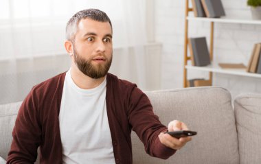 Bir adam elinde uzaktan kumandayla kanepede oturuyor. Rahatlamış ve cihaza odaklanmış görünüyor, tahminen kanal değiştiriyor ya da televizyondaki sesi ayarlıyor..