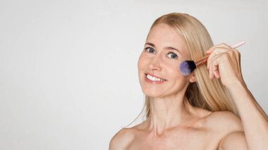 Beyaz bir geçmişi olan sarışın bir kadın bir fırça kullanarak yanağına allık sürüyor. Kameraya bakarken gülümsüyor..