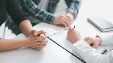 Bu görüntü, tıbbi muayene sırasında bir hastanın ve bir doktorun ellerinin yakın çekimini gösteriyor. Hasta ellerini önlerindeki masanın üzerine yapıştırmış durumda.