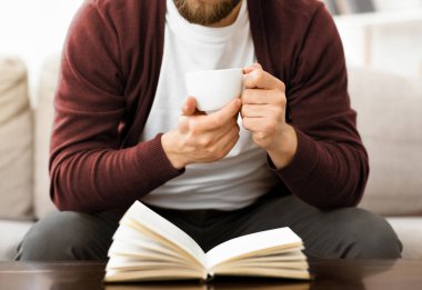 Bir adam bir koltukta oturuyor, elinde bir bardak tutuyor ve bir kitap okumaya dalmış. Rahat ve metne odaklanmış görünüyor, rahat bir duruşu var, kırpılmış