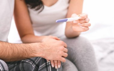 Bir kadın, partnerine hamilelik testi gösterir ve olumlu bir sonuç verir. Çift yatakta oturuyor, ve kadın da testi partnerinin önünde yapıyor, kırpılmış.