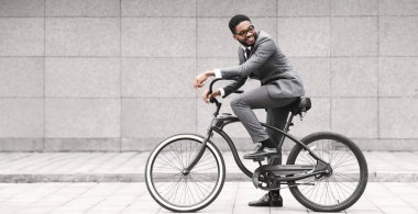 Profesyonel bir siyah adam bisikletine binmiş, şehir merkezinde gri bir duvarın yanında duruyor. Kentsel ve dinamik bir ruh hali, işe gidip gelmeye çağdaş bir yaklaşımı vurguluyor..