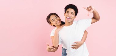 Neşeli bir Afro-Amerikan çifti, pembe bir arka plana karşı neşeli bir şekilde poz veriyor. Kız onu arkadan kucaklarken, erkek şakacı el hareketleriyle gülümsüyor.