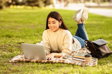 Asyalı kadın çimlere dizüstü bilgisayar kullanarak yaslanıyor. O, klavyeye yazı yazarak işine daldı. Yeşil çimenler etrafını sarmış ve dizüstü bilgisayar yüzünü aydınlatıyor..