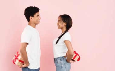 Genç bir siyah erkek ve kız canlı pembe bir arka planda bir arada duruyorlar, ellerinde renkli paketlenmiş hediyeler tutarken gülümsüyorlar, boşluğu kopyalıyorlar.