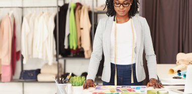 Odaklanmış bir Afrikalı Amerikalı kadın moda tasarımcısı masasında duruyor, önüne serilmiş çeşitli renkli kumaş örneklerini inceliyor, panorama
