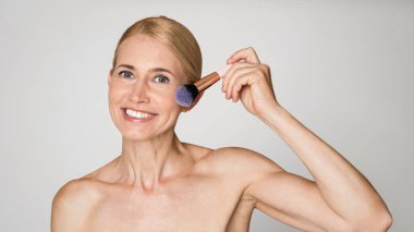 Sarı saçlı olgun bir kadın yanağına allık fırçasıyla makyaj yapıyor. Gülümsüyor ve doğrudan kameraya bakıyor. Arka plan düz beyaz bir duvar..