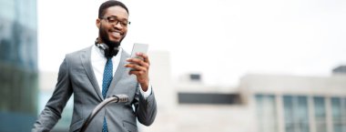 Bisikletli bir Afrikalı Amerikalı işadamı telefonunu kullanıyor. Ortam sakin ama profesyonelce, modern bir yaşam tarzını açık havada yakalıyor..