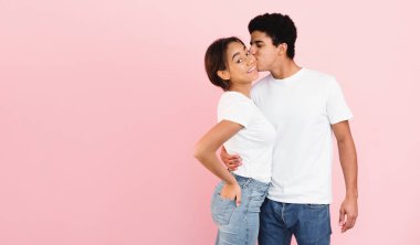 Afro-Amerikalı bir erkek ve kız öpüşürler, parlak pembe bir arka planın önünde dururlar. Çiftlerin kucaklaşması şefkatli ve samimi, aralarında sevgi dolu bir an yaratıyor, kopyalama alanı.