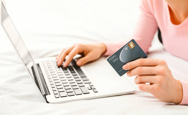 A woman laying on bed, focused on her laptop screen, holding a credit card in her hand. She is likely making an online purchase or checking her financial information, cropped