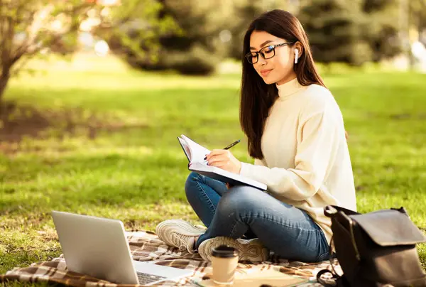 stock image Chinese young woman with glasses sits cross-legged on a checkered blanket in a park. She is studiously writing in a notebook while wearing earphones, with a laptop, a cup of coffee