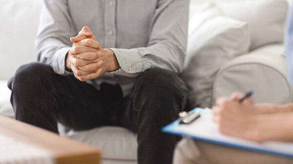 A man sits on a couch with his hands clasped, speaking with a therapist who takes notes on a clipboard during a session in a cozy living room setting, cropped