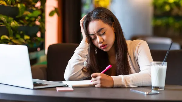 stock image A young black girl is concentrating hard while studying in a cozy cafe. She is surrounded by a laptop, a notebook, and a pen, and appears deep in thought