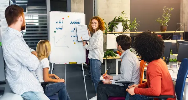 stock image A confident young woman stands presenting a chart to a group of attentive colleagues during a collaborative meeting in a contemporary workspace