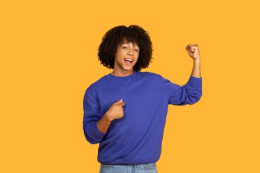 Koyu kıvırcık saçlı ve büyük bir gülümsemesi olan genç bir adam sarı bir arka fonda duruyor, diğer eliyle kendini gösterirken kaslarını esnetiyor. Kendinden emin ve gururlu görünüyor.