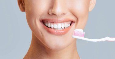 Gülümseyen bir kadın yüzü, diş fırçası ve diş macunu ağzının yanında duruyor. Görüntü onun parlak beyaz dişlerine ve mutlu ifadesine odaklanıyor..