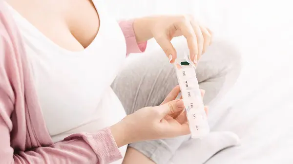 stock image A woman is organizing her daily medication into a pillbox while sitting comfortably on a bed. She is wearing casual, home attire, with a soft focus on her hands and the pillbox, cropped